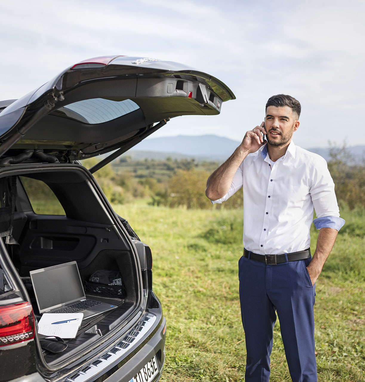 Felix Kuster steht bei geöffnetem Kofferraum an seinem schwarzen Auto und telefoniert mit einer Hand in der Hosentasche.