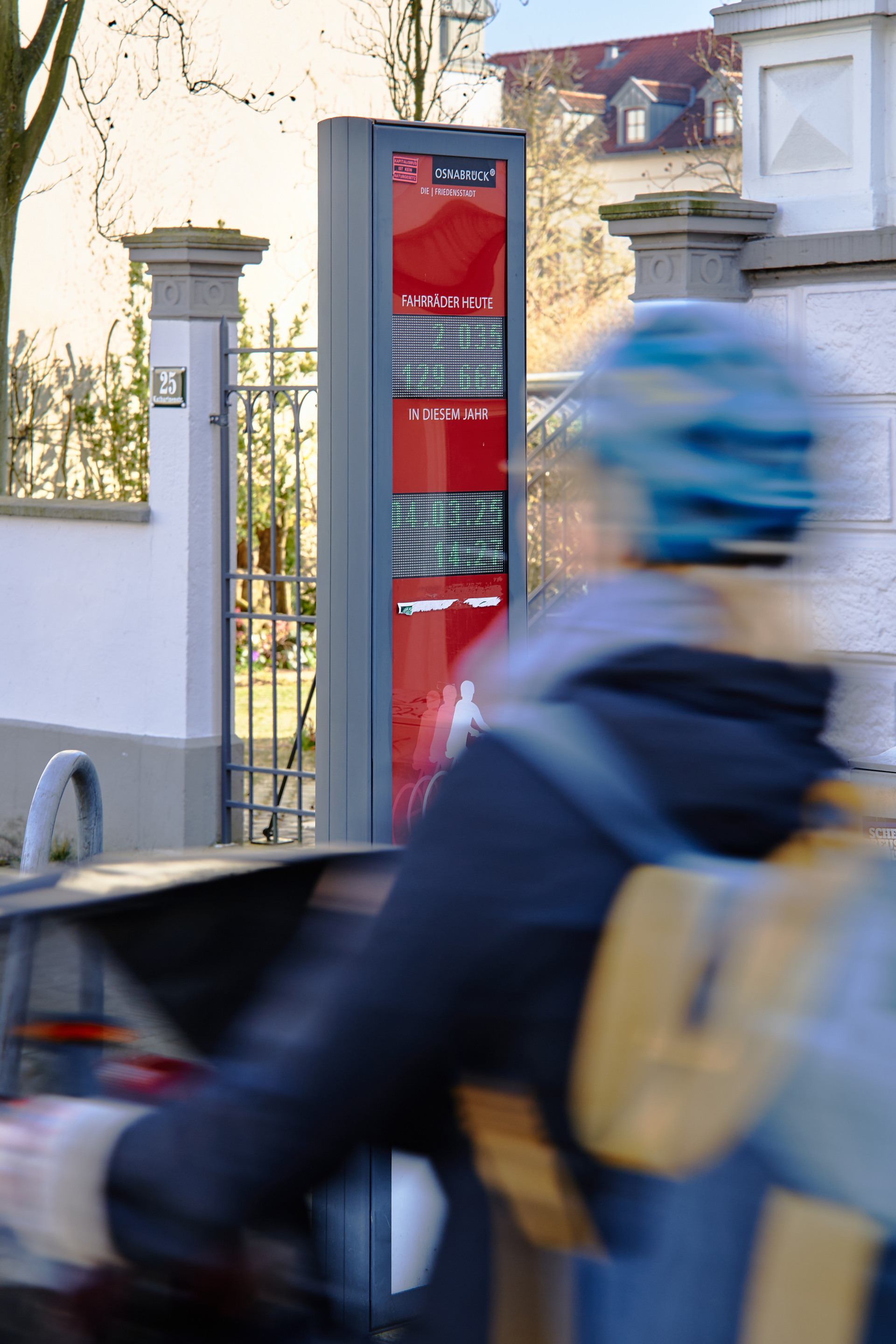 Eine unscharfe Person mit einem blauen Helm, die an einem Informationsstand für öffentliche Verkehrsmittel vorbeigeht. Im Hintergrund ist ein rotes Informationsschild mit einem digitalen Zähler für Fahrräder sichtbar.