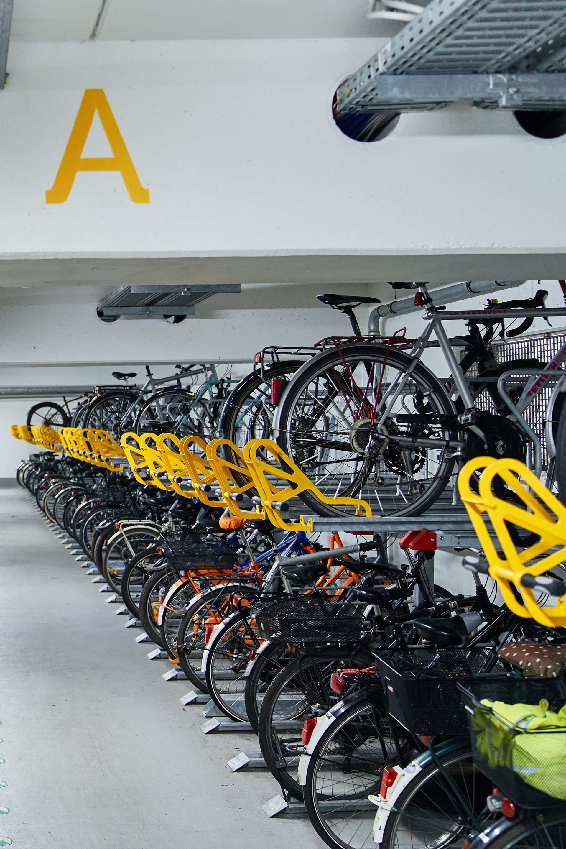 Innenansicht eines modernen Fahrradparks mit mehreren Reihen von Fahrrädern, die auf speziellen Halterungen stehen. Über den Fahrrädern ist ein gelbes 'A' angebracht