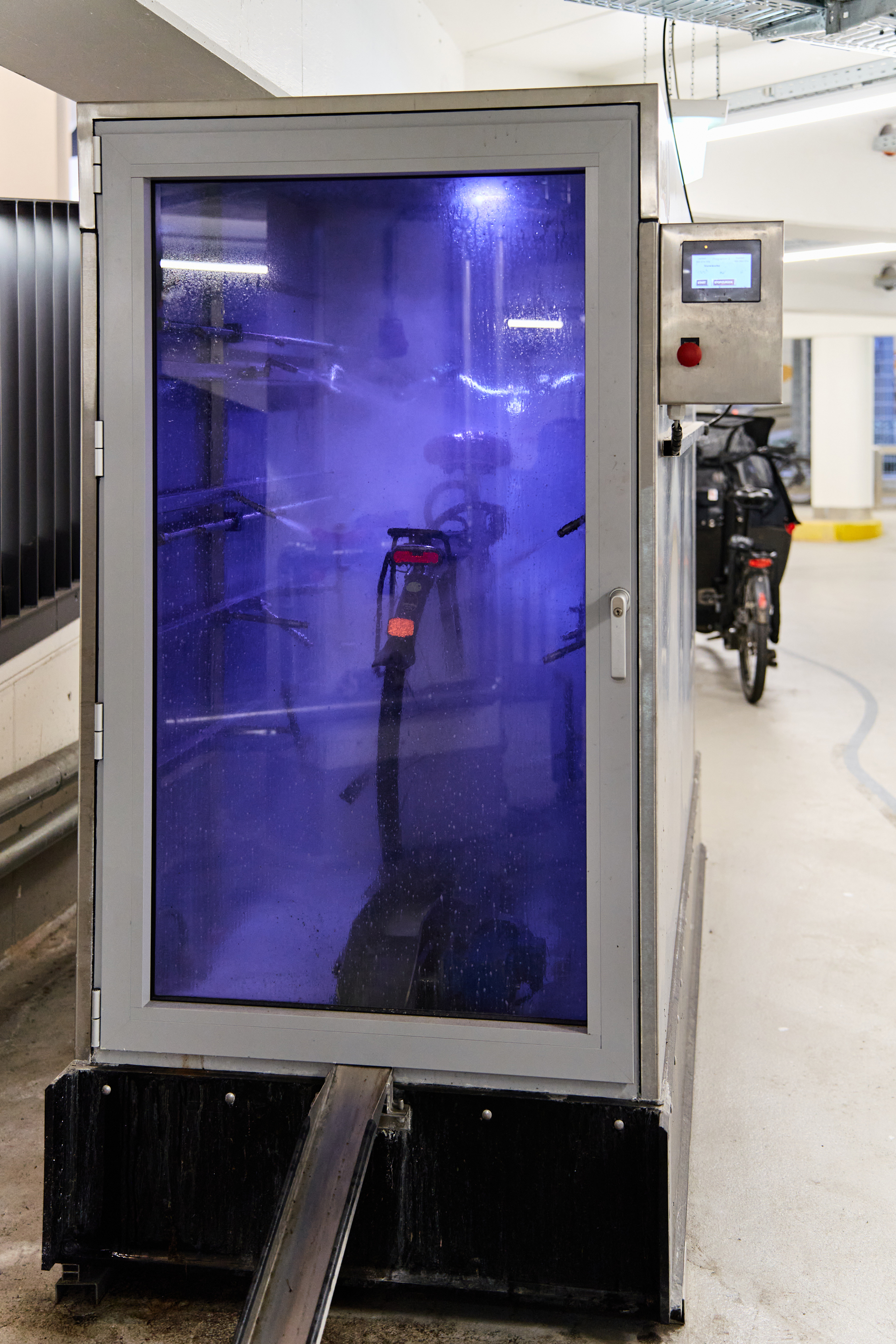 Ein moderner Reinigungsstand für Fahrräder, der mit blauer Beleuchtung und Dampf arbeitet. Im Inneren ist ein Fahrrad sichtbar, während im Hintergrund weitere Fahrräder abgestellt sind.