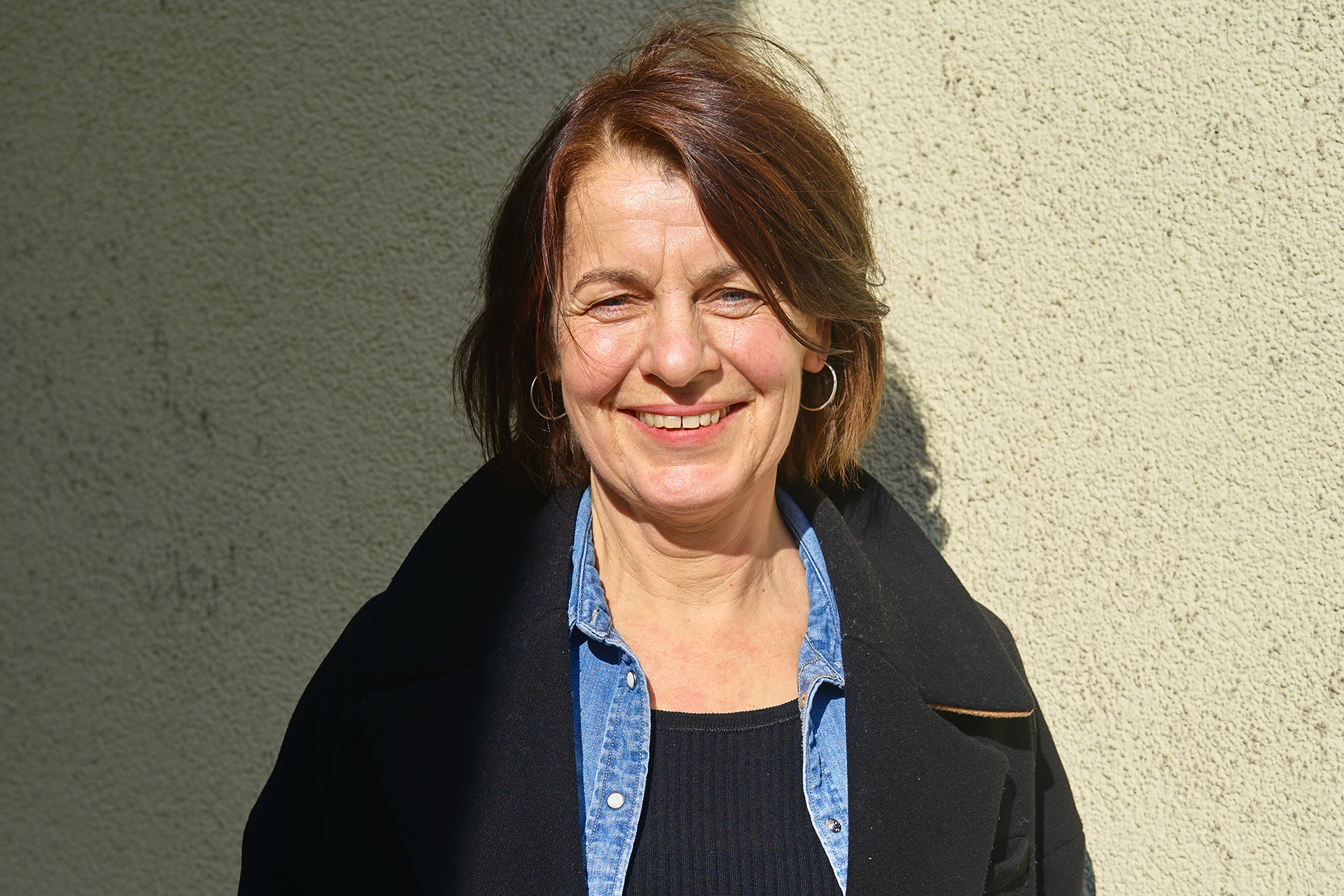 Lächelnde Frau in einem schwarzen Mantel und blauer Bluse steht vor einer hellen Wand.