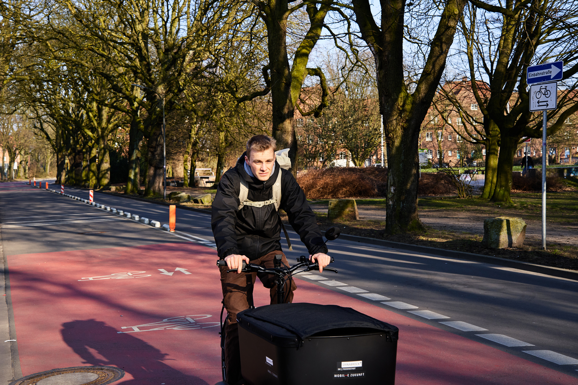 Radfahrer mit einem Lastenrad auf einer Fahrradspur, umgeben von Bäumen und Stadtlandschaft.