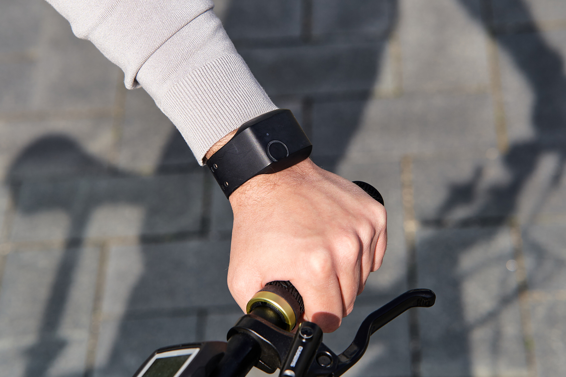 Nahaufnahme einer Hand mit einer Smartwatch, die einen Fahrradlenker hält, auf einem gepflasterten Weg.