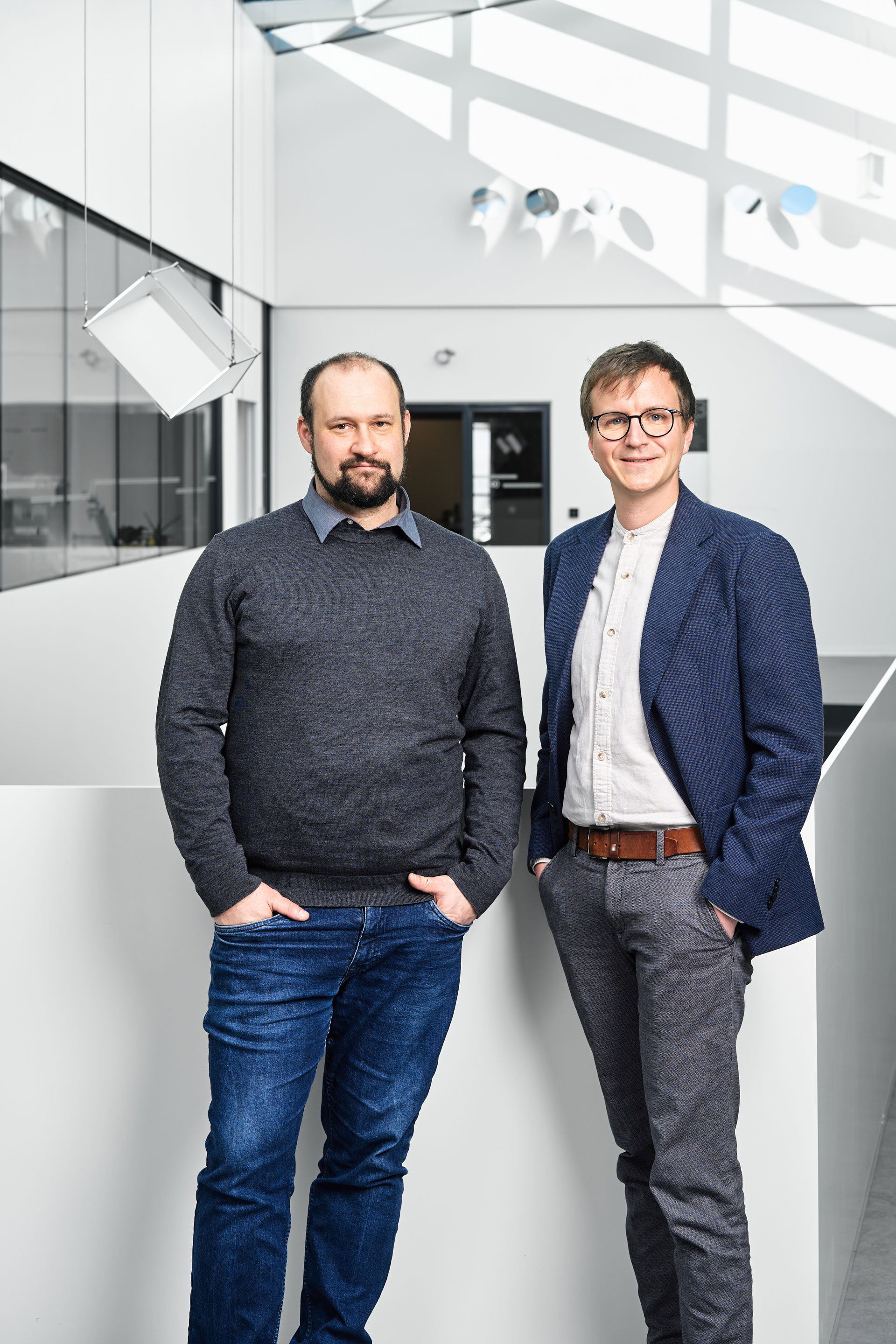 Zwei Männer stehen nebeneinander in einem modernen Büro. Der linke Mann trägt einen grauen Pullover und Jeans, der rechte Mann ist in einem blazer und weißem Hemd gekleidet. Beide blicken freundlich in die Kamera, während im Hintergrund eine helle, minimalistische Architektur sichtbar ist.