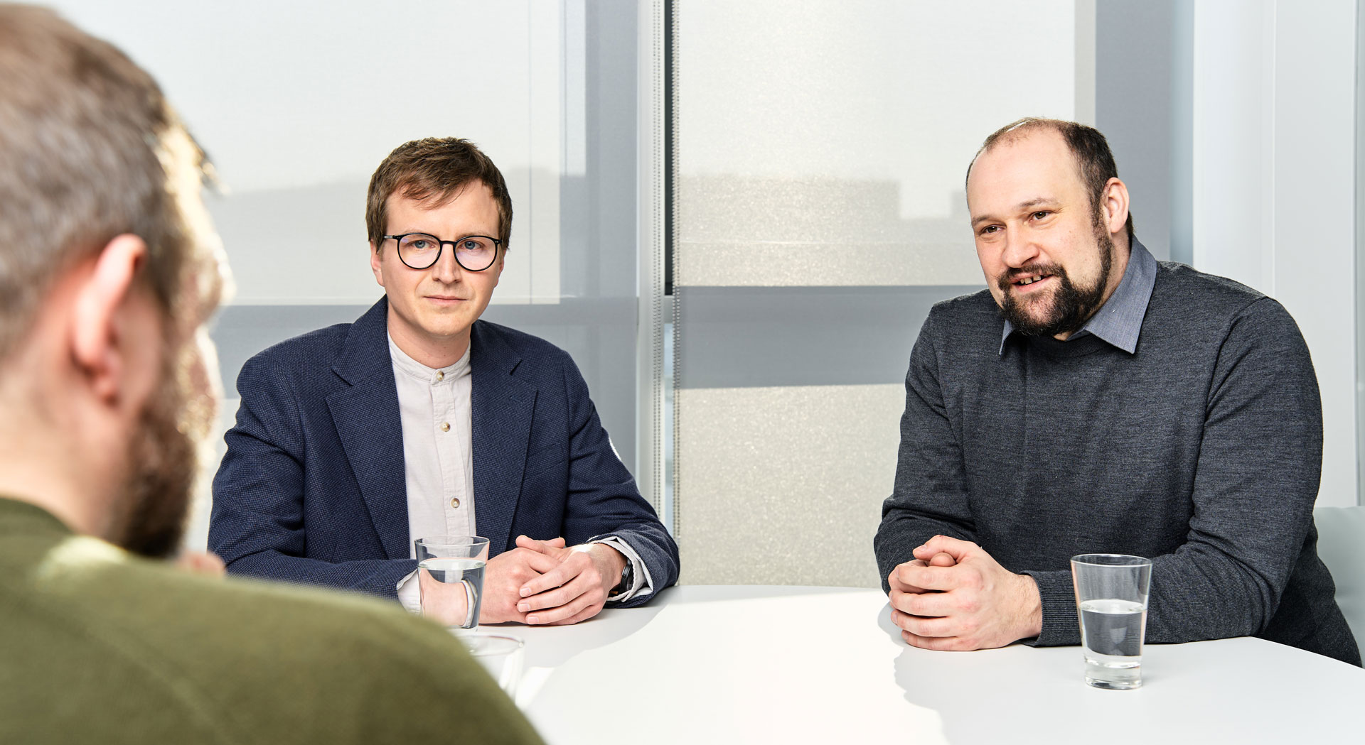 Zwei Personen sitzen an einem Tisch in einem modernen Besprechungsraum. Tobias Höpfer auf der linken Seite trägt eine Brille und einen dunklen Blazer, während Sebastian Stegmüller auf der rechten Seite einen grauen Pullover trägt. Auf dem Tisch steht ein Glas Wasser. Im Vordergrund ist der Rücken einer dritten Person zu sehen, die dem Gespräch gegenübersitzt.