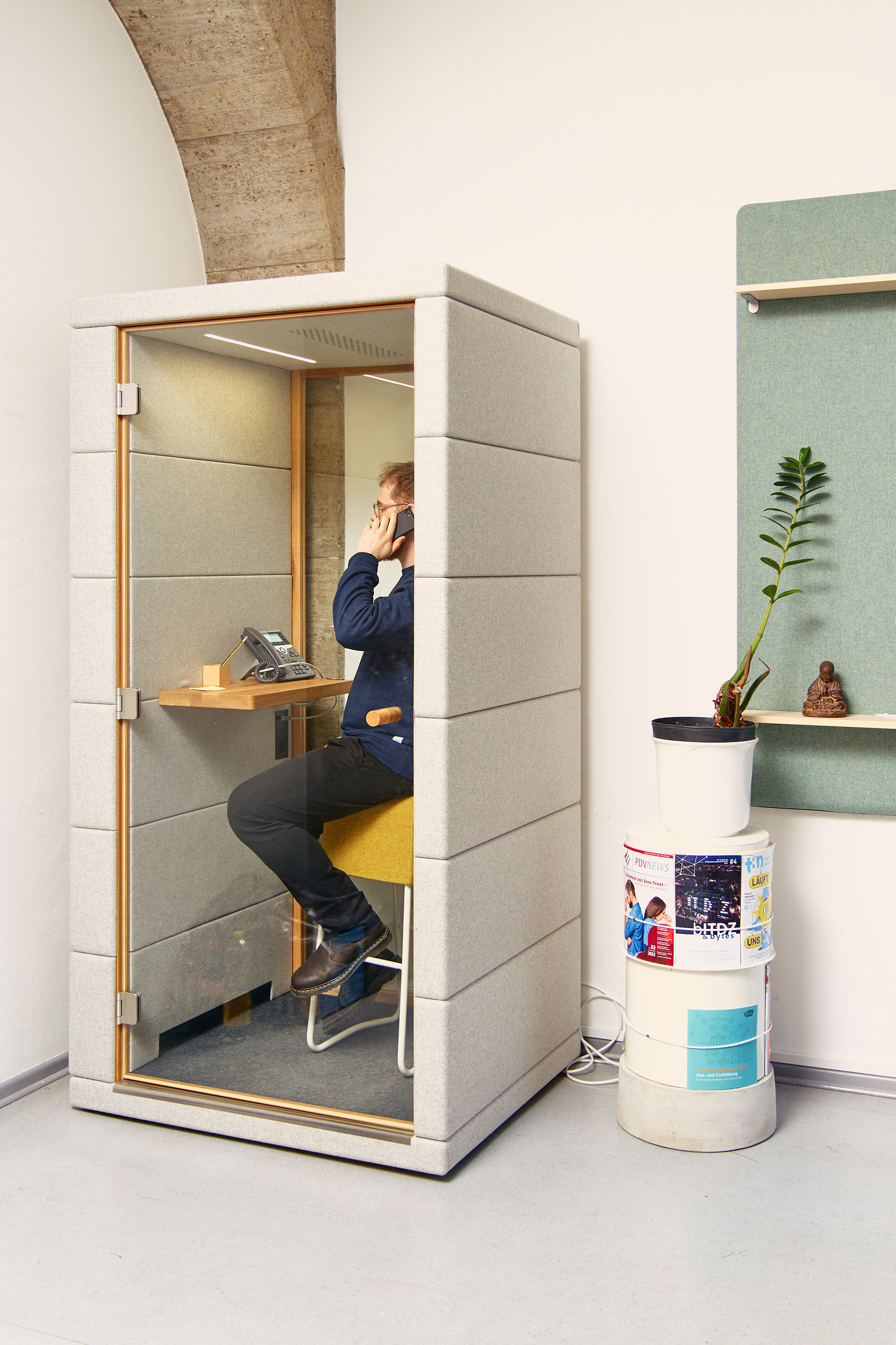 Eine moderne schalldichte Telefonkabine mit grauer Polsterung, in der ein Mitarbeiter auf einem gelben Hocker telefoniert. Die Kabine steht in einem Bürobereich mit historischen Steingewölben.