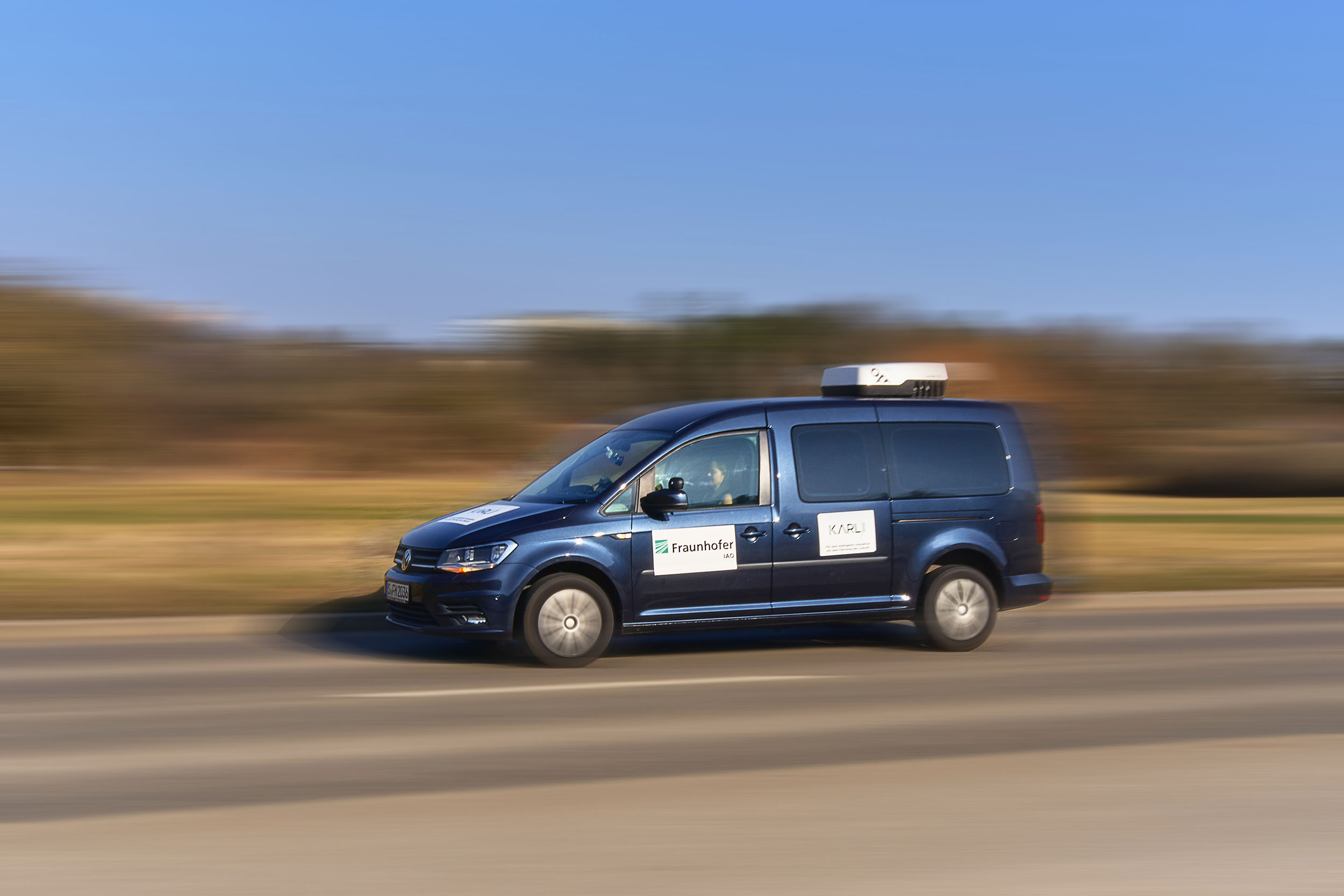 Das Bild zeigt ein Testfahrzeug in Fahrt, aufgenommen in seitlicher Perspektive mit Mitzieheffekt. Auf der Seite des Fahrzeugs sind die Logos der Partner »Fraunhofer IAO« und »KARLI« gut sichtbar. Das Auto fährt auf einer Landstraße bei sonnigem Wetter, der Hintergrund ist bewusst verwischt, um die Bewegung zu betonen. Auf dem Dach befindet sich ein technisches Modul zur Datenerfassung oder Sensorik.