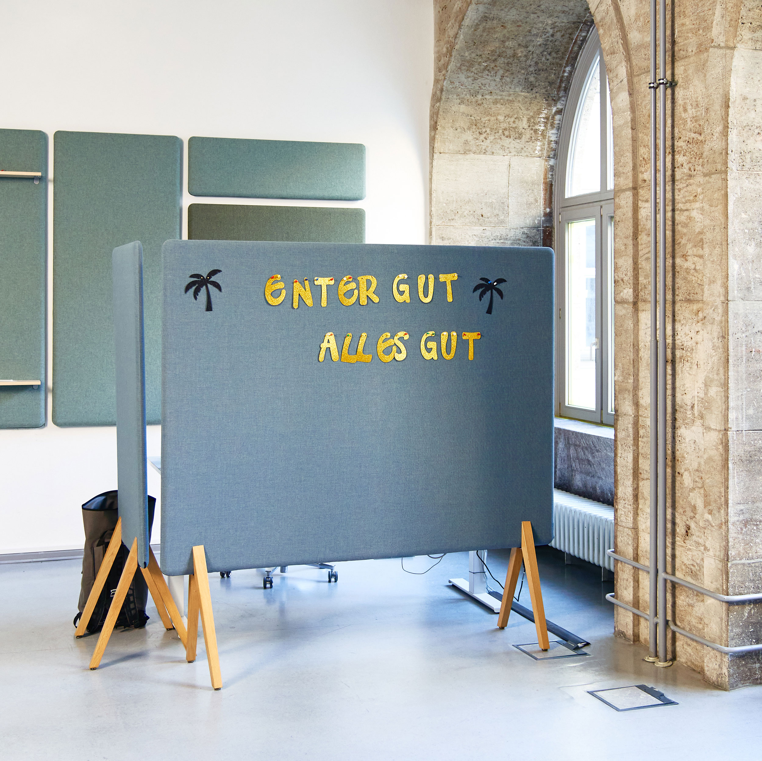 Eine hellblaue mobile Stellwand mit der gelben Aufschrift „ENTER GUT, ALLES GUT“ und kleinen Palmenmotiven steht in einem lichtdurchfluteten Büro zwischen historischen Steinbögen und modernen Akustikpaneelen.