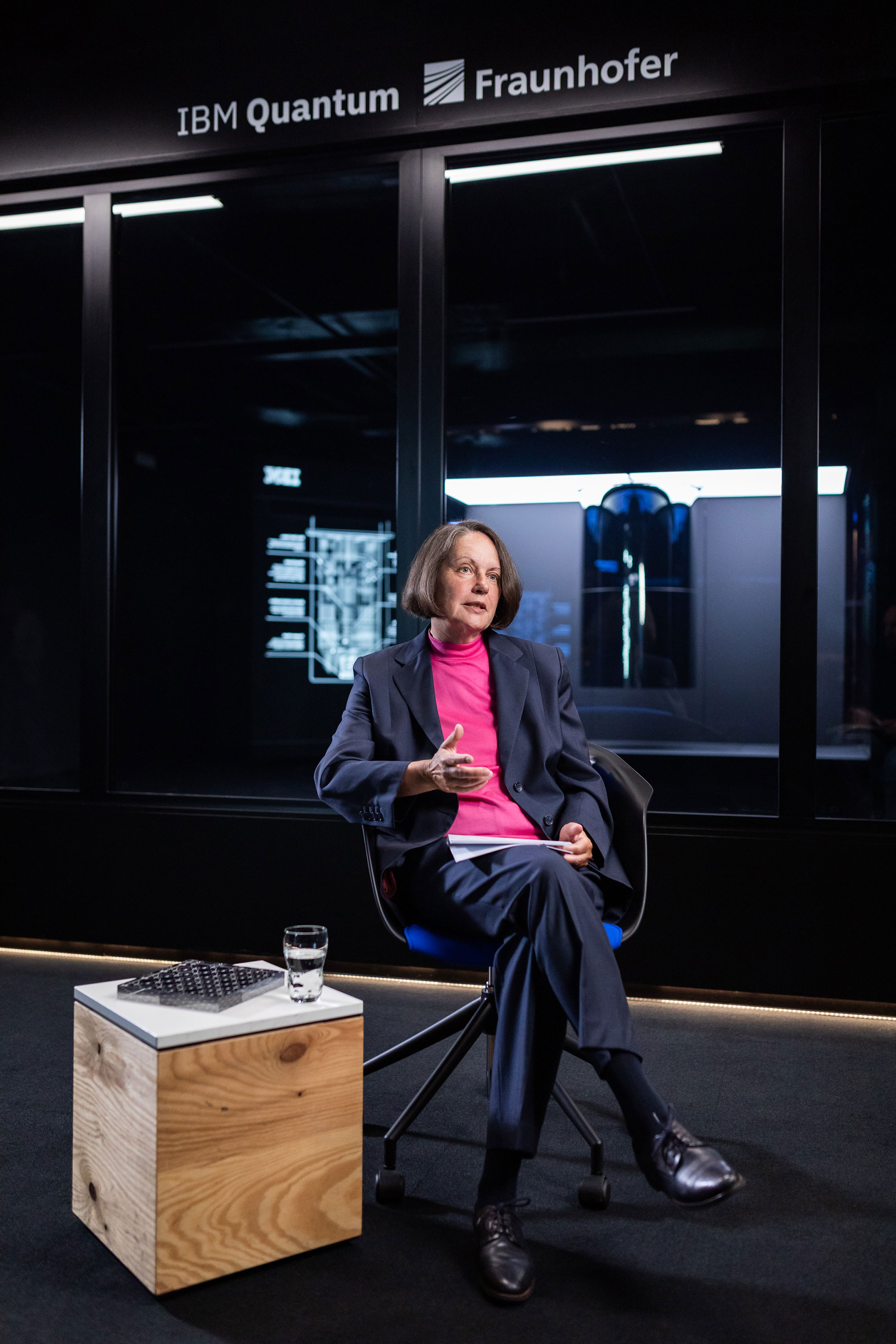 Prof. Dr.-Ing. Anette Weisbecker sitzt auf einem Stuhl und spricht. Im Hintergrund ist der IBM-Quantencomputer zu sehen.