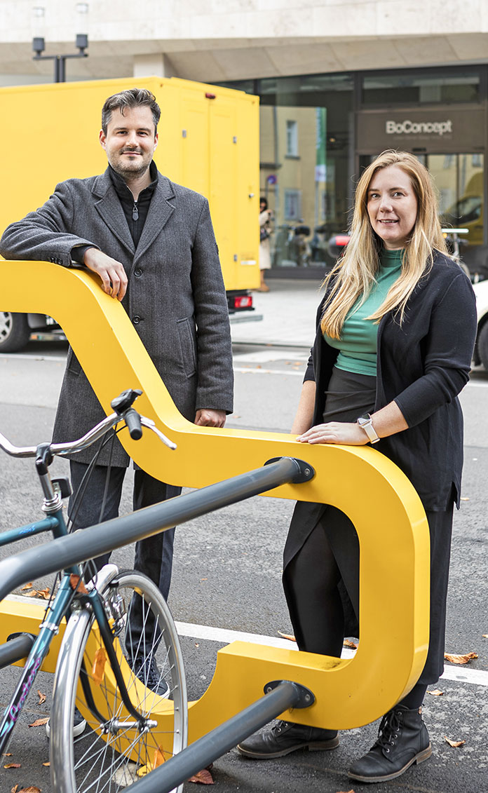 Rebecca Litauer und Lars Mauch, Projektleitende von GreenPickUp stehen nebeneinander an einem gelben Fahrradständer in einer Stadt und sehen lächeln in die Kamera.