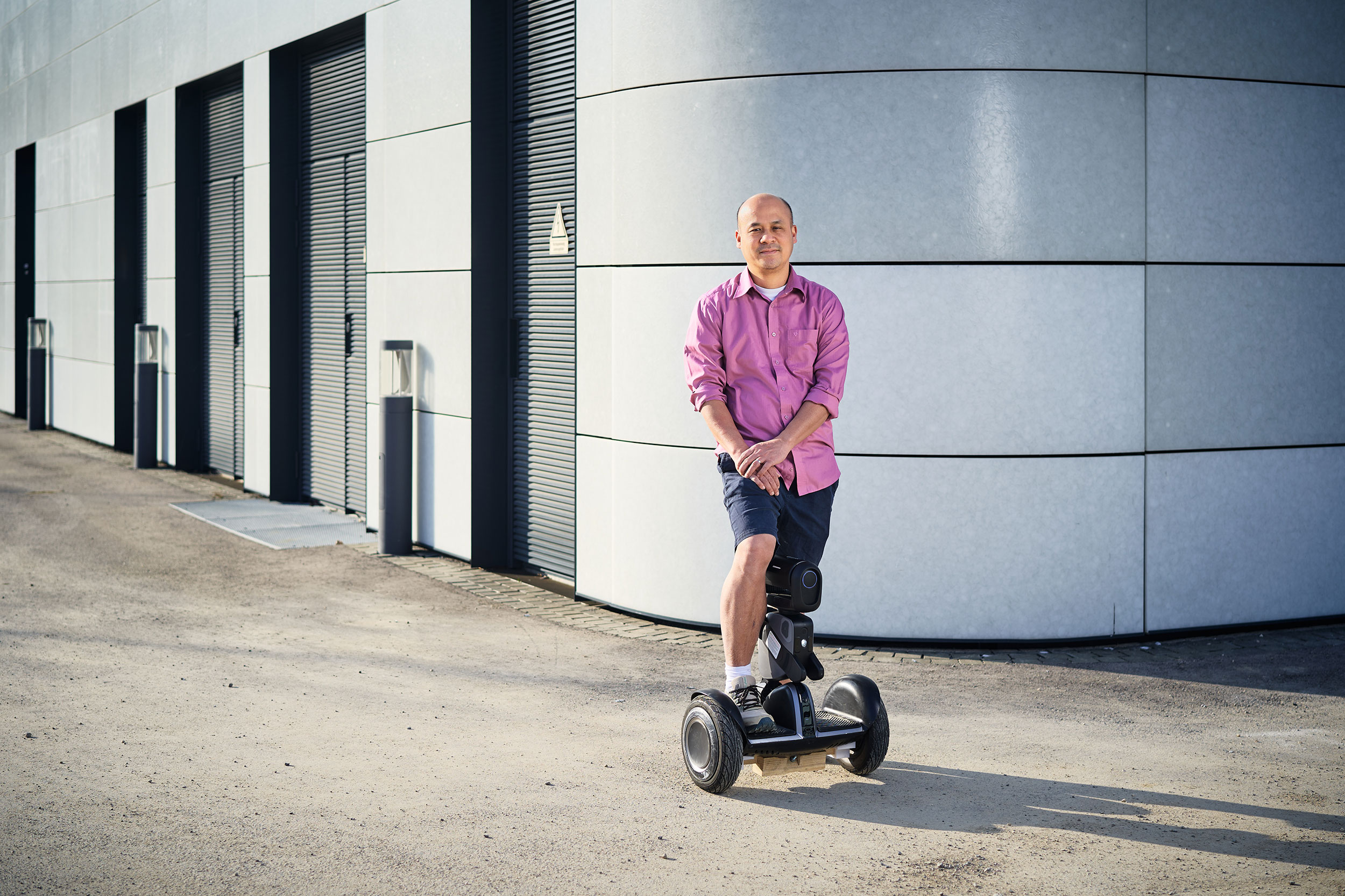 Ein Mann mit kurzen Haaren und einem lila Hemd steht auf einem elektrischen Scooter. Er trägt bermudashorts und hat eine entspannte Haltung, während er mit einem Bein auf dem Scooter steht. Der Hintergrund zeigt eine moderne, graue Wand eines Gebäudes. Das Bild vermittelt ein Gefühl von Mobilität und Innovation.