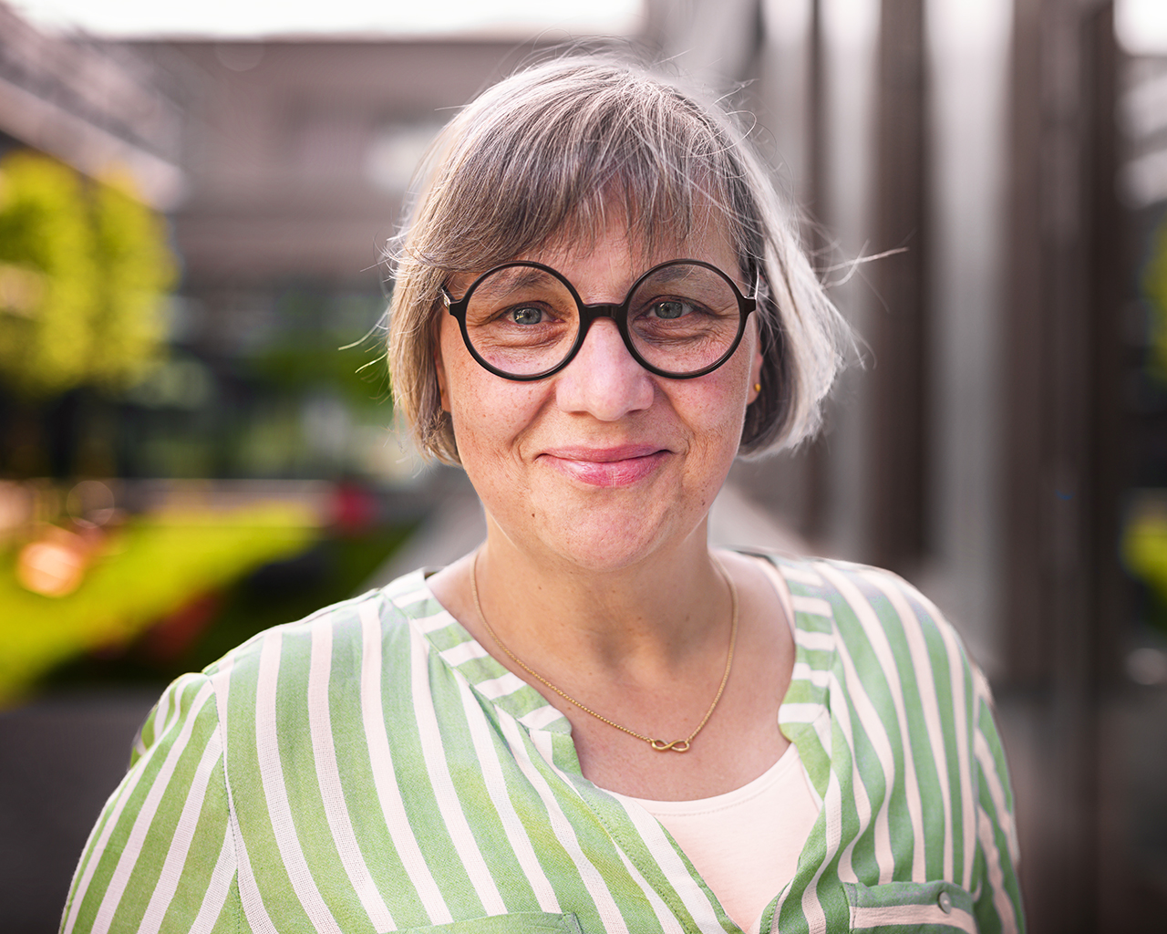 Bild von Petra Holland mit lächelndem Blick in die Kamera. Sie hat einen grauen Bob, trägt eine runde Brille und eine grün-weiß-gestreifte Bluse.