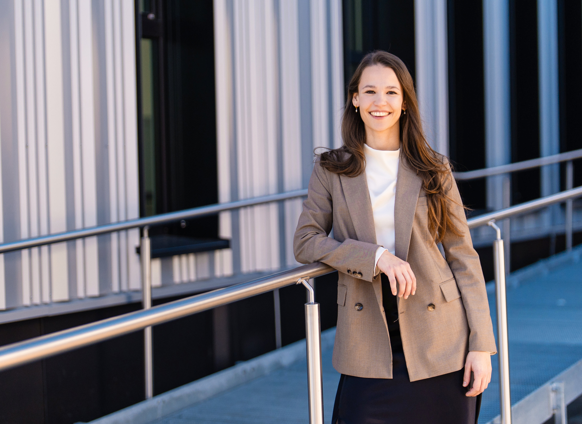 Eine Frau in einem braunen Rock und einem beigen Blazer steht lächelnd an einem Geländer.