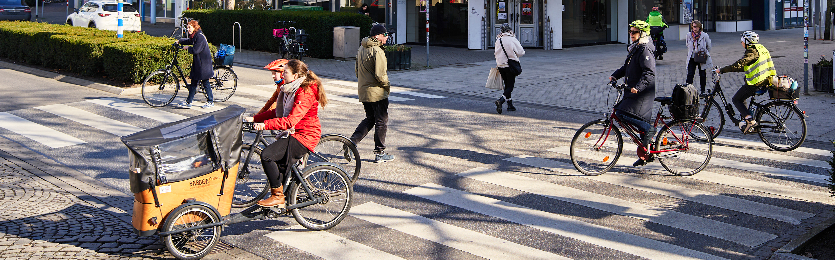 Eine belebte Straßenkreuzung, auf der mehrere Personen auf Fahrrädern unterwegs sind. Im Vordergrund fährt eine Person in einem roten Mantel auf einem Lastenfahrrad. Im Hintergrund sind weitere Radfahrer und Fußgänger zu sehen, die die Straße überqueren. Die Szene spielt sich an einem sonnigen Tag in einer städtischen Umgebung ab.