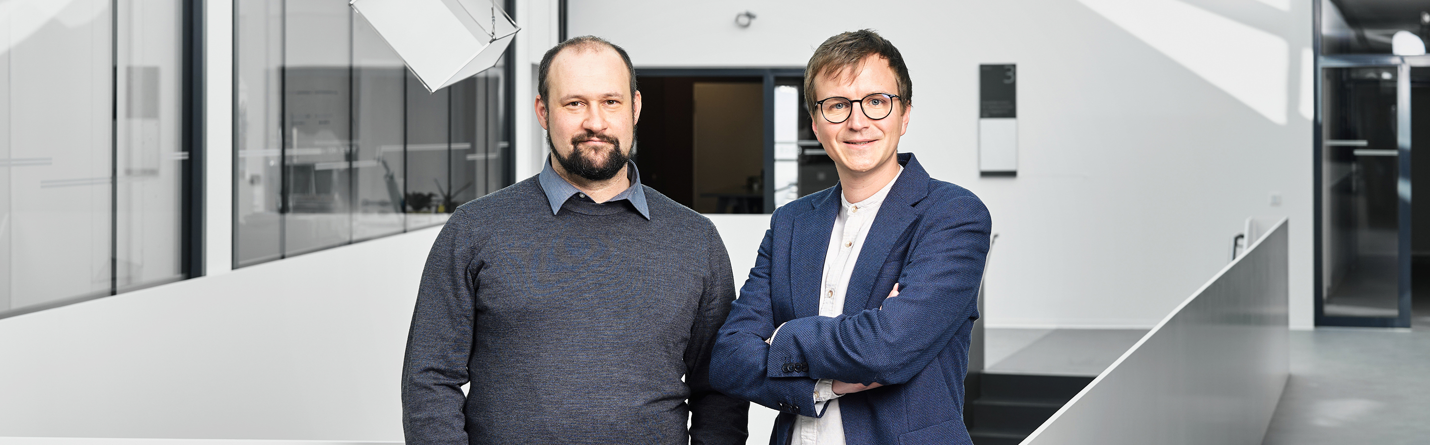Zwei Männer stehen nebeneinander in einem modernen Büro. Der linke Mann trägt einen grauen Pullover und Jeans, der rechte Mann ist in einem blazer und weißem Hemd gekleidet. Beide blicken freundlich in die Kamera, während im Hintergrund eine helle, minimalistische Architektur sichtbar ist.