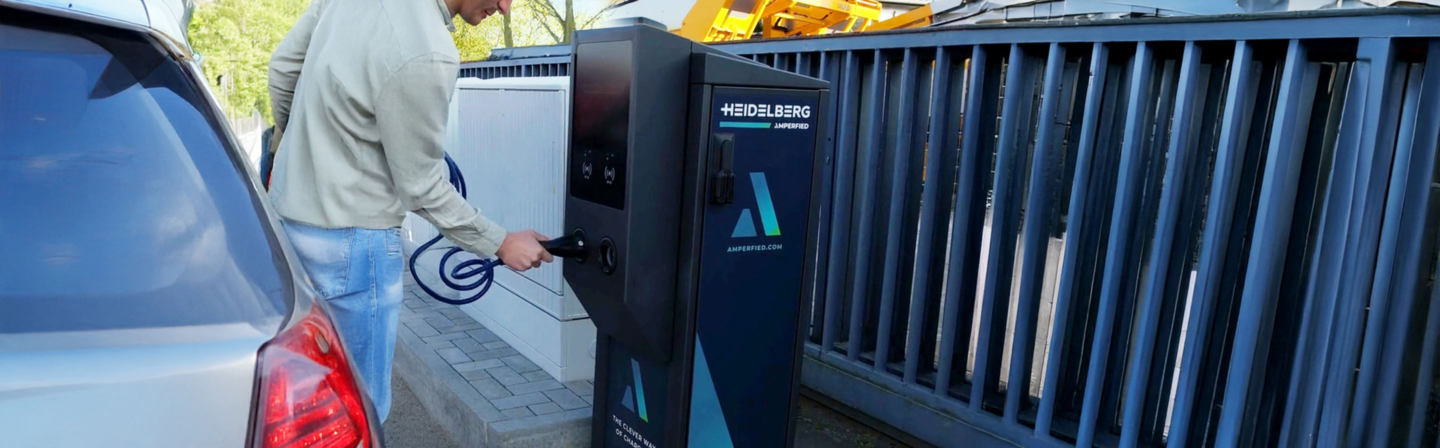 Das Bild zeigt einen Mann, der an einer modernen Ladesäule für Elektrofahrzeuge steht. Er hält das Ladekabel in der Hand und bereitet sich darauf vor, es in das Ladegerät des Autos zu stecken. Im Hintergrund sind weitere Ladesäulen und eine gepflegte Umgebung zu sehen, die auf eine stationäre Ladestation hinweist.