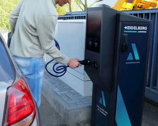 Das Bild zeigt einen Mann, der an einer modernen Ladesäule für Elektrofahrzeuge steht. Er hält das Ladekabel in der Hand und bereitet sich darauf vor, es in das Ladegerät des Autos zu stecken. Im Hintergrund sind weitere Ladesäulen und eine gepflegte Umgebung zu sehen, die auf eine stationäre Ladestation hinweist.