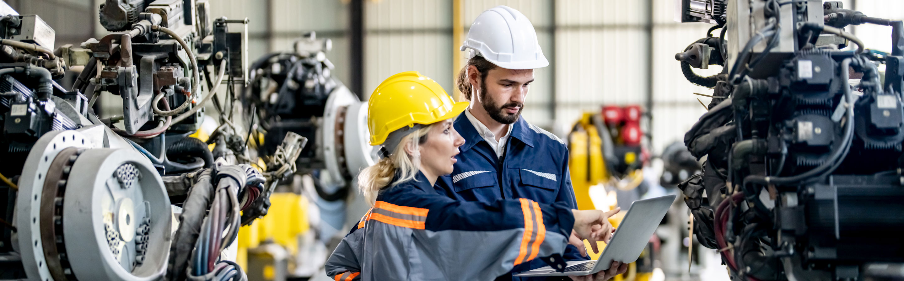 Zwei Fachleute in Schutzkleidung und Helmen arbeiten in einer Industriehalle an einem Laptop, umgeben von großen Maschinen und Robotern.