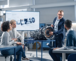 In einem modernen Techniklabor präsentiert ein Ingenieur einer kleinen Gruppe ein großes Motorteil; Bildschirme im Hintergrund zeigen „Industry 4.0“.