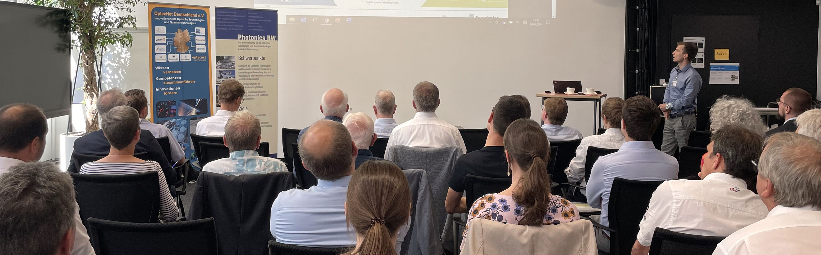 Ein Publikum beobachtet einen Vortrag und blicken auf eine Leinwand.