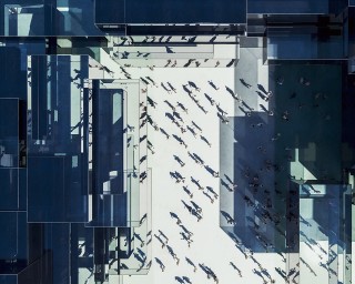 zukunft-handel-innenstadt-teaser Vogelperspektive auf eine belebte Stadtlandschaft mit zahlreichen Menschen, die sich zwischen modernen, transparenten Gebäuden bewegen. Der Boden ist hell und reflektiert das Licht, während die Schatten der Gebäude eine interessante Musterung erzeugen.