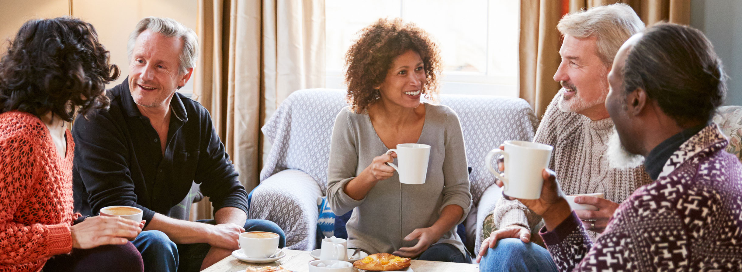 Fünf Personen, darunter drei männliche Rentner und zwei jüngere Frauen, sitzen zusammen, trinken Kaffee und unterhalten sich.