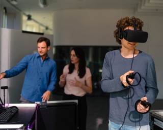 Mehrere Personen sind abgebildet; eine Person agiert mit einer VR-Brille und Controllern, während die anderen beiden am Computer sitzen und Daten einsehen.